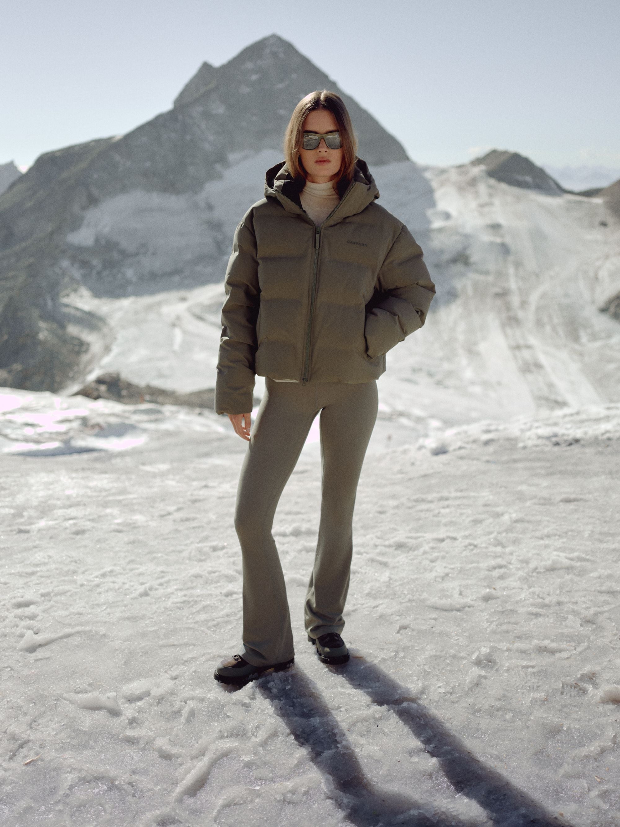 Woman wearing a khaki CASPARA Aurora puffer jacket in a snowy mountain landscape.